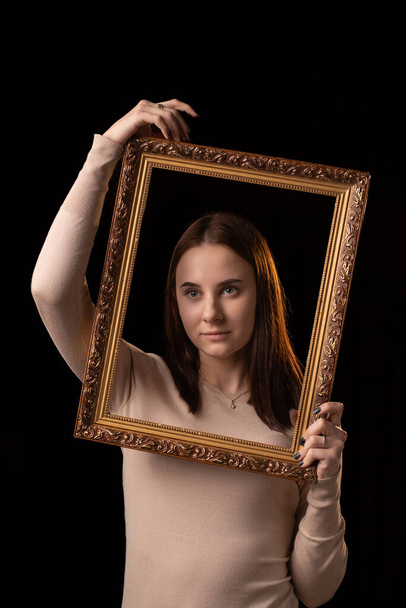 A young woman is holding a picture frame. Black background. Psychological portrait, introspection, reflection concept. - Foto, imagen