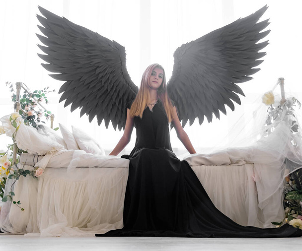 An angel with black wings on a white bed in flowers. A young thoughtful girl is sitting on the edge of the bed. - Foto, imagen