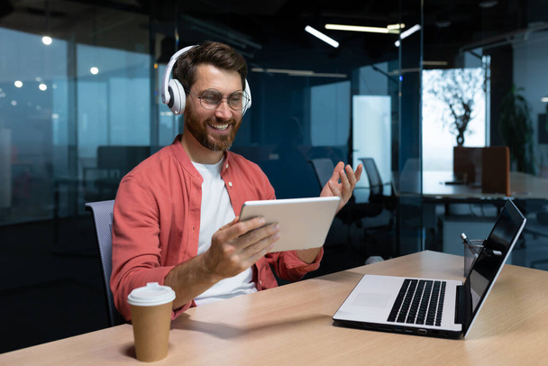 A young businessman freelancer, student, programmer sits in the office at the table with a laptop in headphones, holds and uses a tablet. Talks on a video call, listens to music. - Foto, Imagen