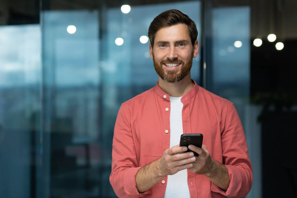 Portrait of a young man in a red shirt standing in the office, holding a phone in his hands, looking at the camera with a smile. - Foto, afbeelding