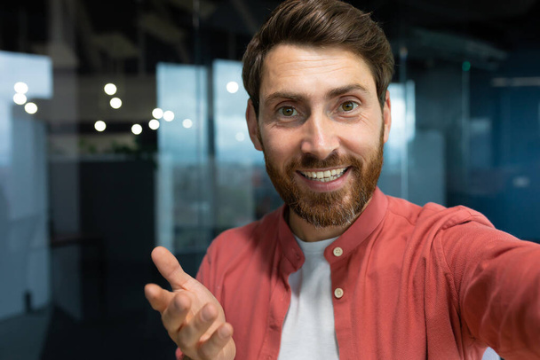 Close-up photo. Young businessman, freelancer, blogger in a red shirt in the office talking on a video call from the phone, smiling at the camera. - Zdjęcie, obraz