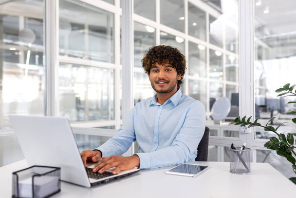 Portrait of young programmer inside office with laptop, indian man smiling and looking at camera, man typing on keyboard, writing code for software, happy with achievement results. - Foto, imagen
