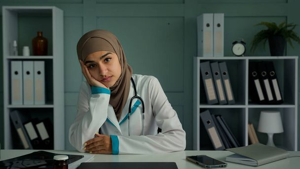 Znudzony nieszczęśliwy przemyślany kobieta muzułmanin lekarz stażysta siedzieć w klinice biurowej czuć smutek zaburzenia psychiczne myśleć depresja myśli leniwy lekarz bez motywacji zmęczony po pandemii koronawirusa - Zdjęcie, obraz