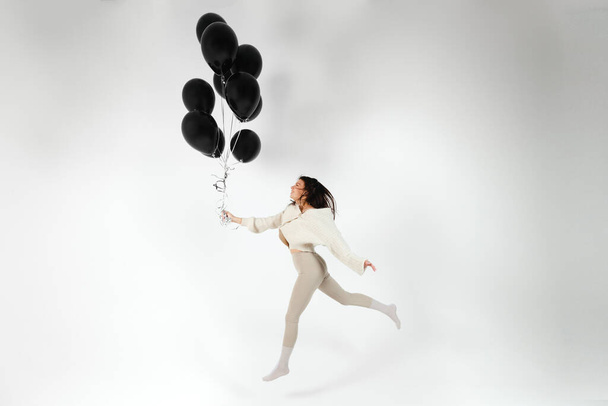 Bruneta dívka v lehkém oblečení skákání s hromadou černých balónků. Izolováno na bílém pozadí. - Fotografie, Obrázek
