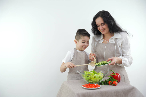 mom and son preparing salad identical beige aprons vegetables cucumbers are poured into a glass plate smiling having a good time with children parent-child relationship caring for the family - Fotoğraf, Görsel