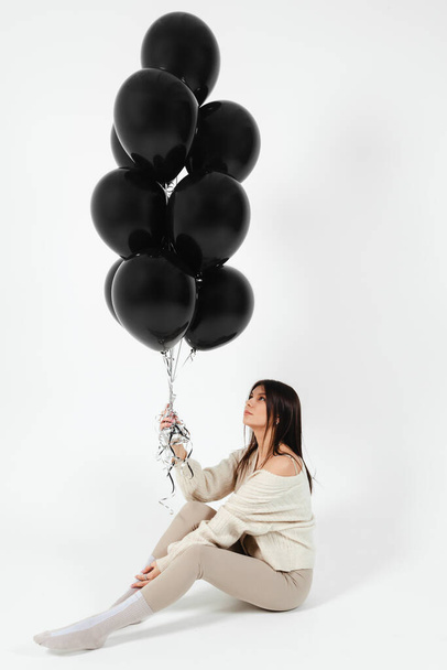 Brunette Girl in light clothes sitting on a floor and a Bunch of black balloons pushes her up. - Фото, изображение