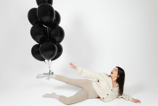 Açık renk elbiseli esmer kız yerde yatıyor ve bir grup siyah balon onu bacağından yukarı itiyor.. - Fotoğraf, Görsel