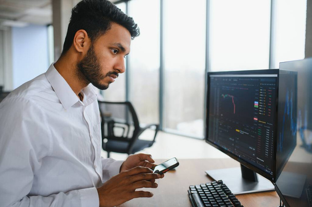Side view of smart Indian crypto trader, investor, analyst broker, using laptop and smartphone analyzing digital cryptocurrency exchange, stock market charts, thinking of investing and funds risks - Photo, image