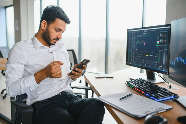 Side view of smart Indian crypto trader, investor, analyst broker, using laptop and smartphone analyzing digital cryptocurrency exchange, stock market charts, thinking of investing and funds risks. - Valokuva, kuva