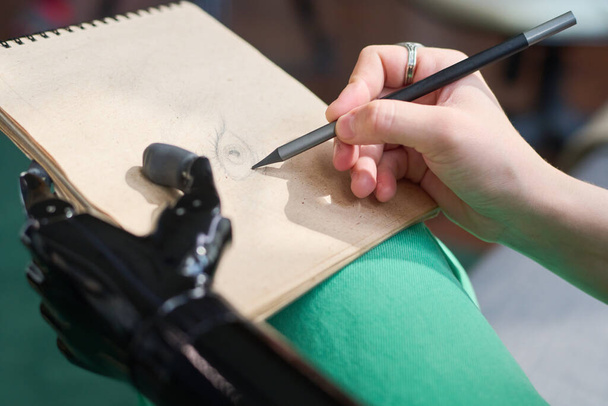 Рука молодого креативного дизайнера с миоэлектрическим протезом рисует глаза на странице блокнота, проводя досуг дома - Фото, изображение