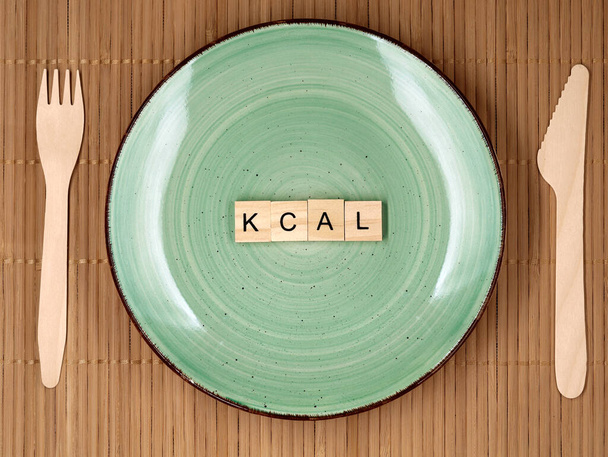 木製のナイフとフォーク、あまりにも多くのカロリーを食べるの食事の概念イメージと緑のプレート上のkcal単語と木製の文字ブロックのトップビュー. - 写真・画像