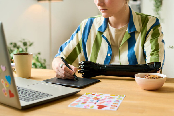 Νεαρός γραφίστας με γραφιστική και tablet που ρετουσάρει εικόνες ή ζωγραφίζει ενώ κάθεται δίπλα στο γραφείο μπροστά από το laptop στο γραφείο του σπιτιού - Φωτογραφία, εικόνα