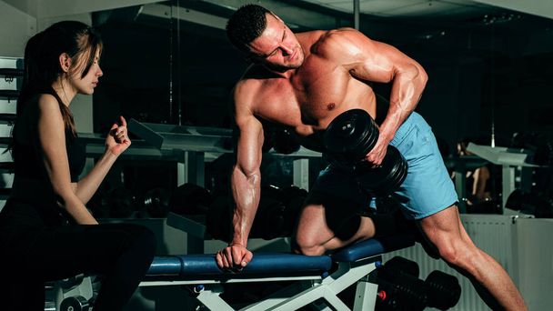 Sporty couple. Woman trainer with muscular man doing functional training workout doing biceps lifting dumbbell. Sportsman doing exercises with weights - 写真・画像