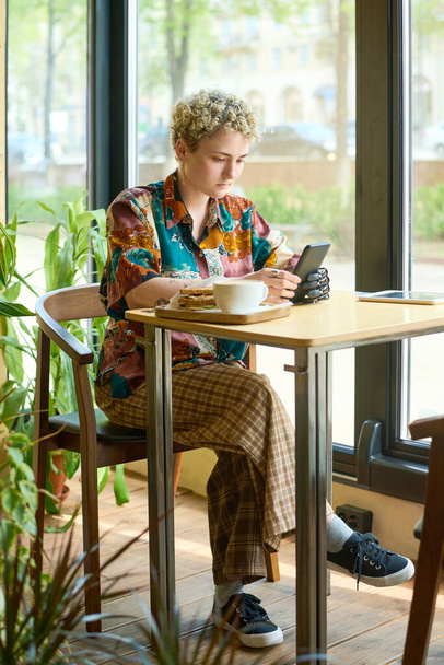 Młoda kobieta w ubraniu casualwear trzymająca smartfona myoelektryczną ręką podczas oglądania wideo online lub komunikowania się w czacie wideo w kawiarni - Zdjęcie, obraz