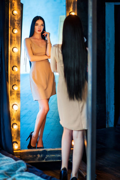 Portrait of a beautiful slim long-haired brunette caucasian girl in beige dress on high heels attractively posing in front of full-length mirror with bulbs looking at own reflection  - Zdjęcie, obraz
