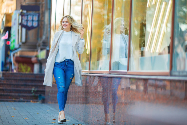 Horizontales Porträt einer schönen jungen kaukasischen Blondine, die lächelnd in der Nähe der Schaufensterscheibe spaziert und ihre Haare fixiert, blaue Jeans und beiger Trenchcoat, verschwommener blaugelber Herbsthintergrund - Foto, Bild