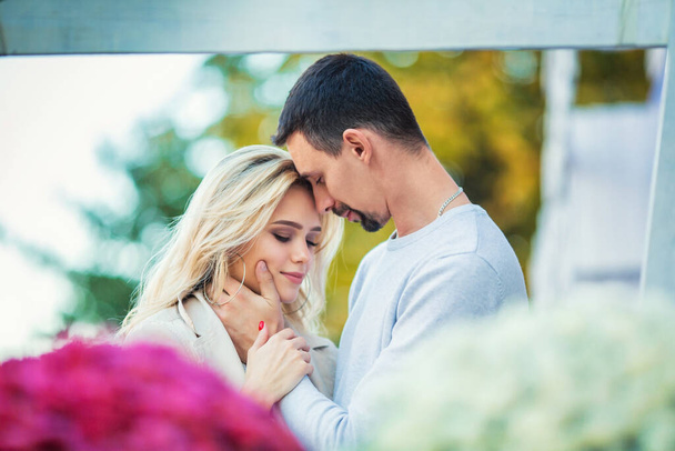 Love story engagement session of a happy young caucasian straight couple in white sweaters, boyfriend is hugging girlfriend, emotionally holding her face, flowers foreground, blurred autumn background - Foto, Imagen