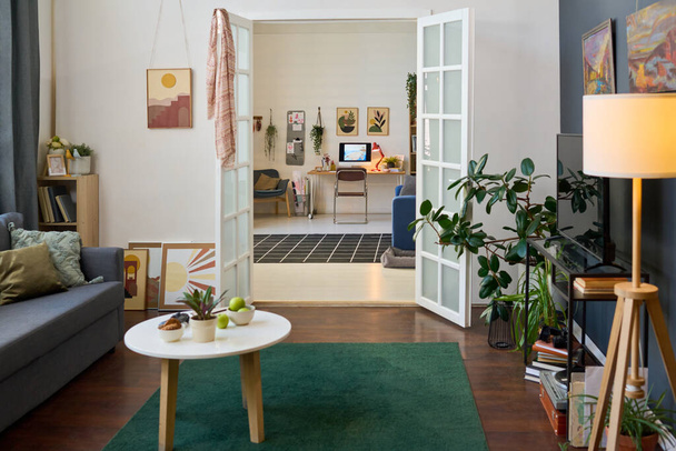 Interiér prostorného obývacího pokoje s otevřenými dveřmi do další velké místnosti s pracovištěm, pohovkou, křeslem a židlí v moderním bytě - Fotografie, Obrázek