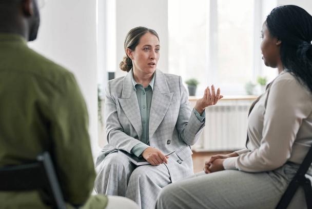 Moderne jonge psychotherapeut communiceert met Afro-Amerikaanse vrouwelijke patiënt terwijl ze voor haar zit tijdens de sessie - Foto, afbeelding