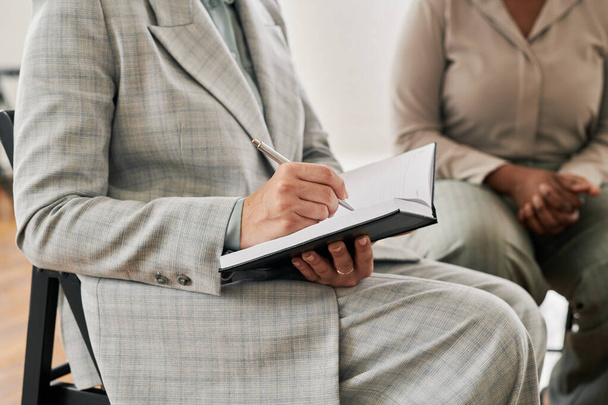 Psicoanalista mujer en traje gris sosteniendo pluma sobre página de cuaderno abierto mientras escribe quejas de pacientes en sesión - Foto, Imagen