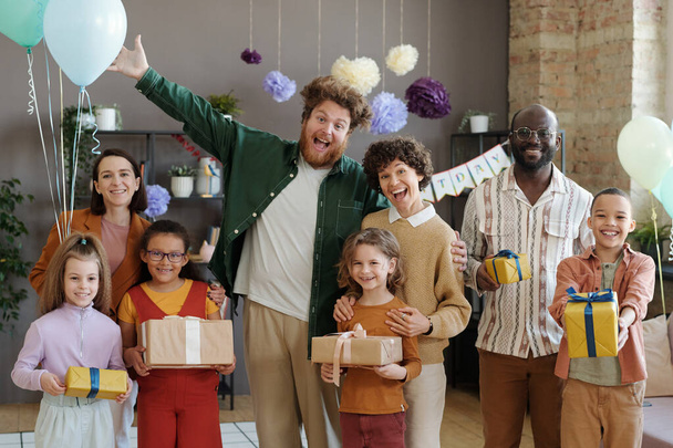 Porträt aufgeregter Eltern und ihrer glücklichen Freunde mit Kindern, die ihren Geburtstag mit Geschenken und Luftballons feiern, sie stehen lächelnd in die Kamera - Foto, Bild