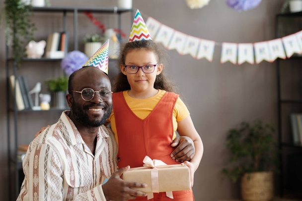 Porträt eines glücklichen afrikanischen Vaters, der zusammen mit seiner Tochter in die Kamera lächelt und ihr ein Geschenk macht, sie tragen im Urlaub Partyhüte - Foto, Bild