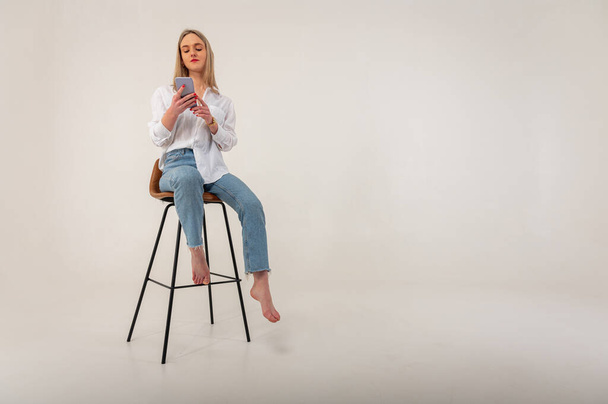 Full length side profile foto van jong aantrekkelijk meisje in jeans en wit shirt kijken, browsen mobiele telefoon terwijl zitten op kruk geïsoleerd op witte achtergrond - Foto, afbeelding
