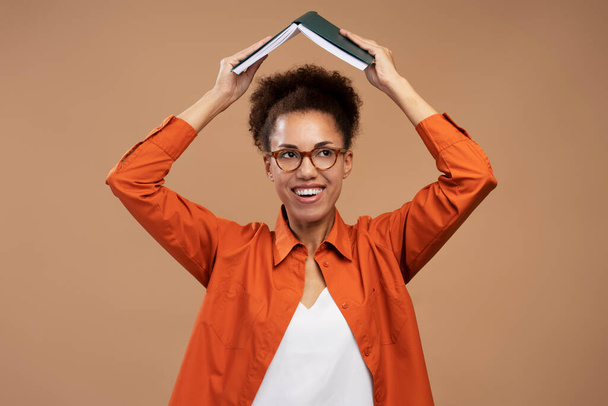 Portrait of positive woman with black hair in eyeglasses, standing holding book over head, studying and education. Indoor studio shot isolated on brown background  - Foto, Bild