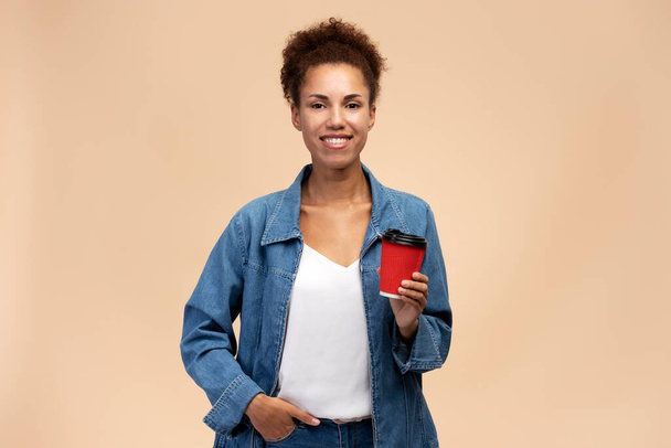 Beautiful woman holding coffee to go and looking to camera, recommend cafe or coffee house. Indoor studio shot isolated on beige background  - Fotoğraf, Görsel