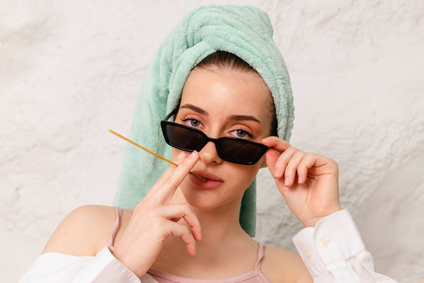 Close up Portrait of a pretty girl with big green eyes wearing black sunglasses, with a towel wrapped around her head and holding a straw in her hand like a cigarette. - Foto, Bild