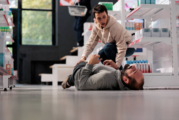 Woman running to man having seizure in medical retail store. Pharmacy scared customer rushing to give first aid to person lying on floor suffering from epileptic attack and foaming at mouth - Fotografie, Obrázek