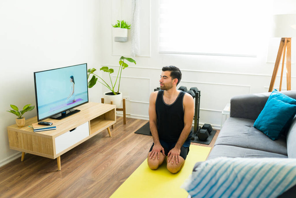 Joven hispano viendo un video de yoga en la televisión y haciendo una pose de rayo durante su entrenamiento en casa - Foto, imagen