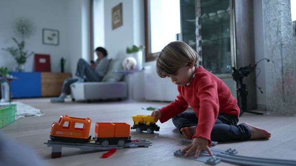 Παιδί παίζει με το τρένο παιχνίδι στο σύγχρονο σπίτι σαλόνι πάτωμα. Το αγοράκι παίζει μόνο του με παιχνίδια δώρων. - Φωτογραφία, εικόνα