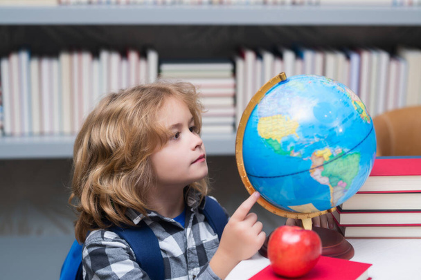 Uczeń patrzący na globus w bibliotece w podstawówce. Dzieciak ze szkoły kujonów. Sprytne dziecko z podstawówki z książką. Mądry geniusz, gotowy do nauki. Globus światowy - Zdjęcie, obraz