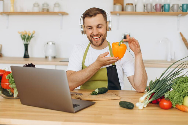 Homem feliz em fones de ouvido sentado na mesa da cozinha e preparando salada, segurando pimenta e mostrando-o ao laptop. - Foto, Imagem