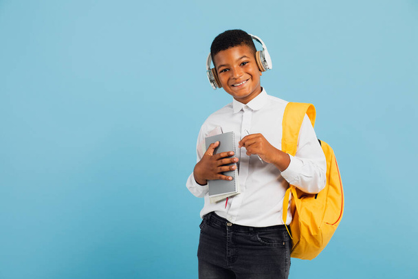 Menino africano feliz usando fones de ouvido e mochila segurando livros e cadernos. Voltar ao conceito de escola. - Foto, Imagem