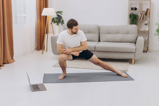 Positive athletic bearded middle-aged man stretching at home, using laptop, watching sport videos on Internet, having fitness class online, copy space. Healthy lifestyle, sport on self-isolation - Photo, image