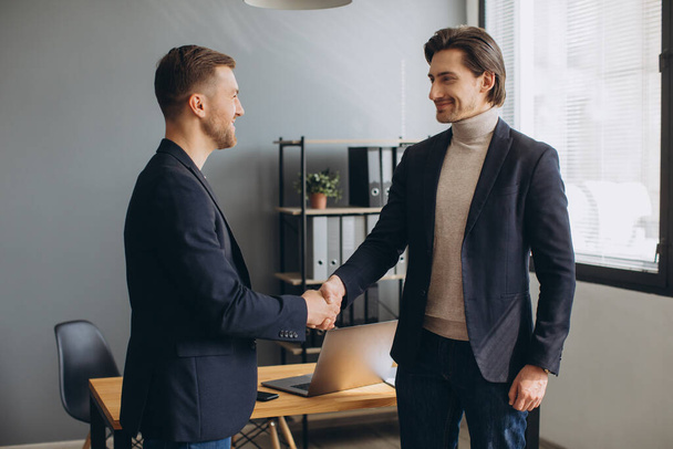 Twee lachende succesvolle jonge zakenmannen schudden elkaar de hand op een zakelijke bijeenkomst in het kantoor - Foto, afbeelding