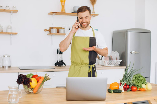 Attractive young man is cooking in the kitchen with a laptop on the table, talking on a smartphone. - Photo, Image