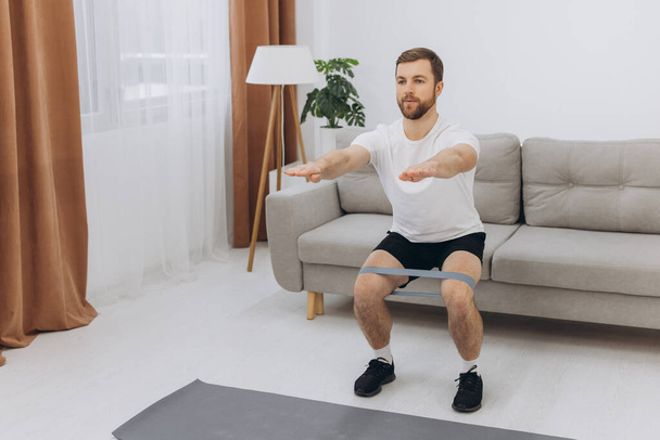 Positive athletic bearded middle-aged man stretching at home, using laptop, watching sport videos on Internet, having fitness class online, copy space. Healthy lifestyle, sport on self-isolation - Photo, Image