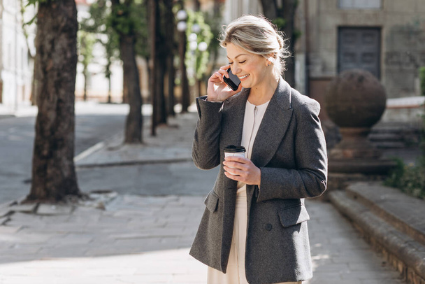 Mature blonde souriante femme d'affaires marchant autour de la ville, buvant du café et parlant au téléphone au printemps sur un fond de fleurs lilas - Photo, image