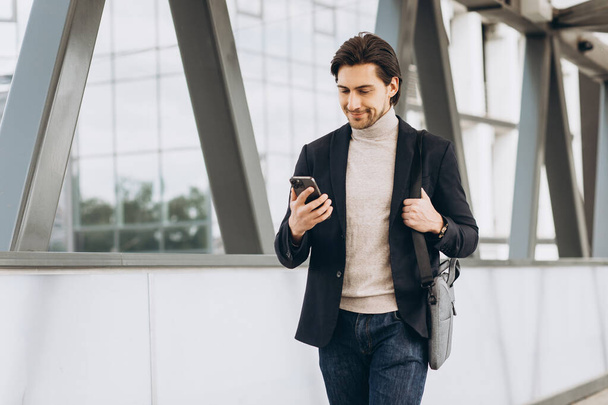 Portrét pohledného moderního mužského podnikatele v obleku chůze s úsměvem a čtení nebo psaní vzkazu na telefonu na pozadí městských budov a kanceláří. - Fotografie, Obrázek