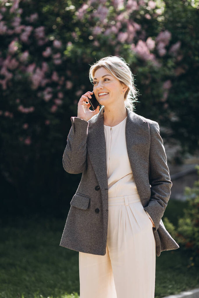 Mulher de negócios loira madura sorrindo andando no parque e falando ao telefone na primavera em um fundo de flores lilás - Foto, Imagem