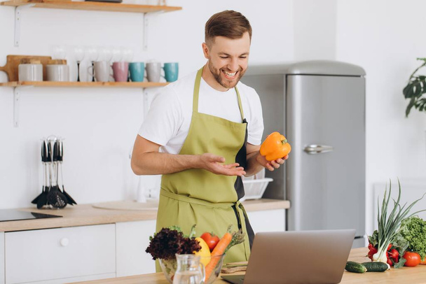 Retrato del hombre feliz en casa, hombre cocinando ensalada de verduras mirando a la cámara y sonriendo, rebanando verduras, utilizando el ordenador portátil para el entrenamiento de cocina en línea - Foto, imagen
