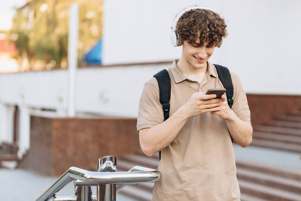 Attractive curly young man university or college student with phone walking around campus - Фото, зображення