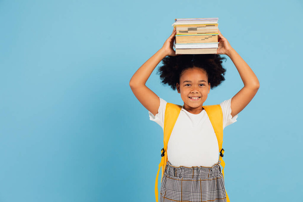 Happy African American schoolgirl holding books above head on blue background, back to school concept. - Photo, image