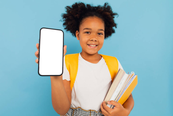 Podekscytowany Little African American School Girl Holding Big Cell in Hand Presenting Telefon pusty ekran na tle Blue Studio. Smartphone Wyświetlacz Mockup - Zdjęcie, obraz