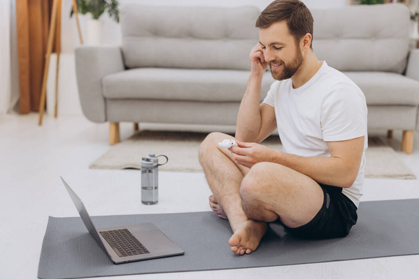 Atrakcyjny brodaty mężczyzna trzymający słuchawki bezprzewodowe w pobliżu laptopa i rozpoczynający poranne ćwiczenia lub jogę. - Zdjęcie, obraz