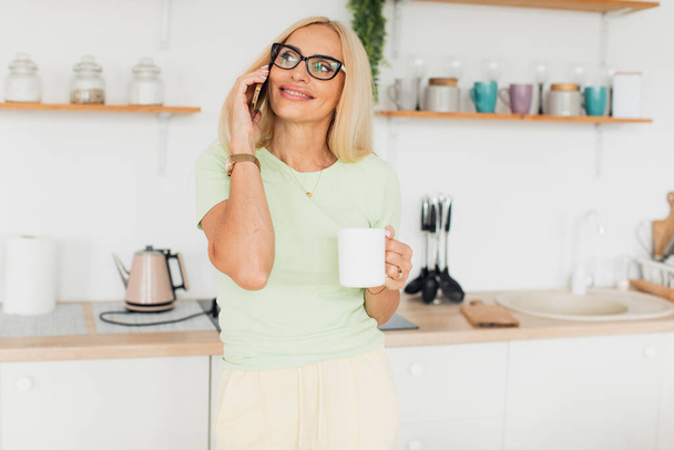 Moderne aantrekkelijke vrouw van middelbare leeftijd praten aan de telefoon en het drinken van koffie in de keuken thuis - Foto, afbeelding