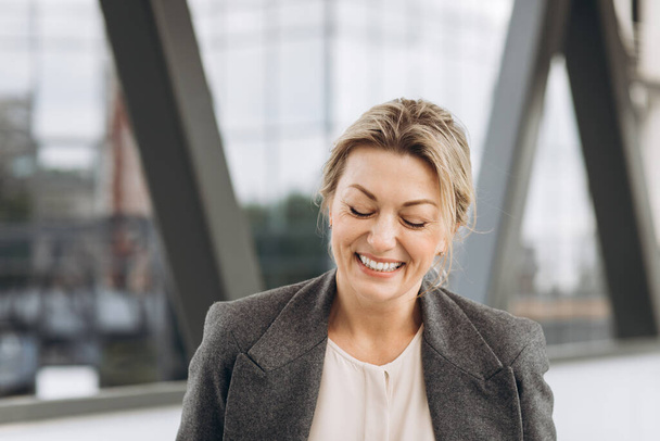 Porträt einer reifen Geschäftsfrau, die vor dem Hintergrund moderner Stadt- und Bürogebäude mit Emotionen lächelt - Foto, Bild